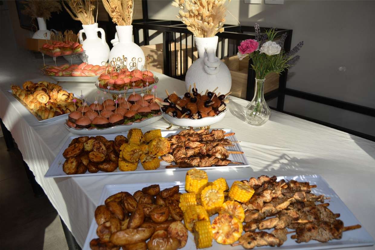 A long buffet table of grilled skewers, roast potatoes, corn and sliders, dressed with white vases and fresh flowers
